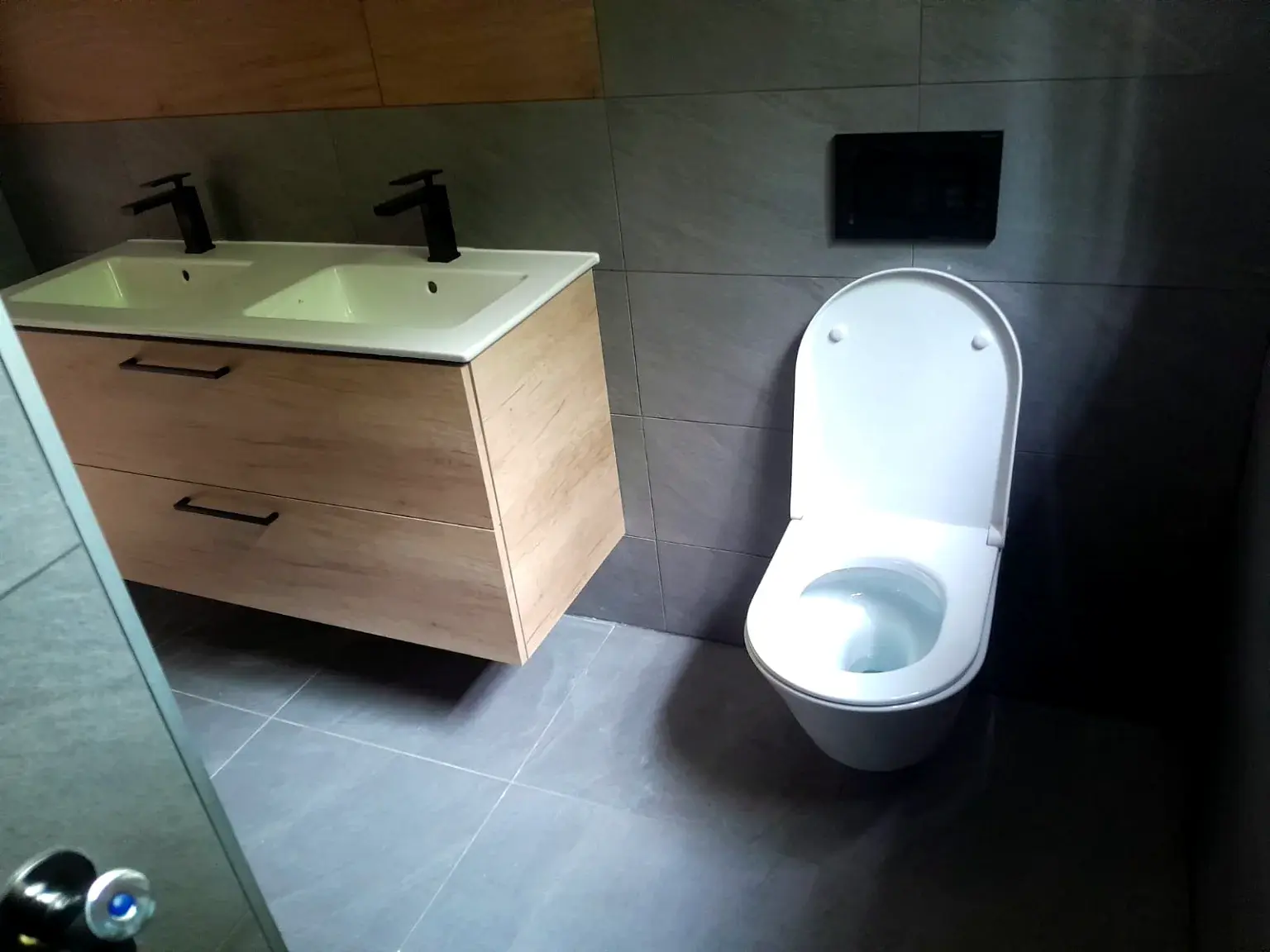 Salle de bains sombre avec double vasque bois clair et sanitaires suspendus.
