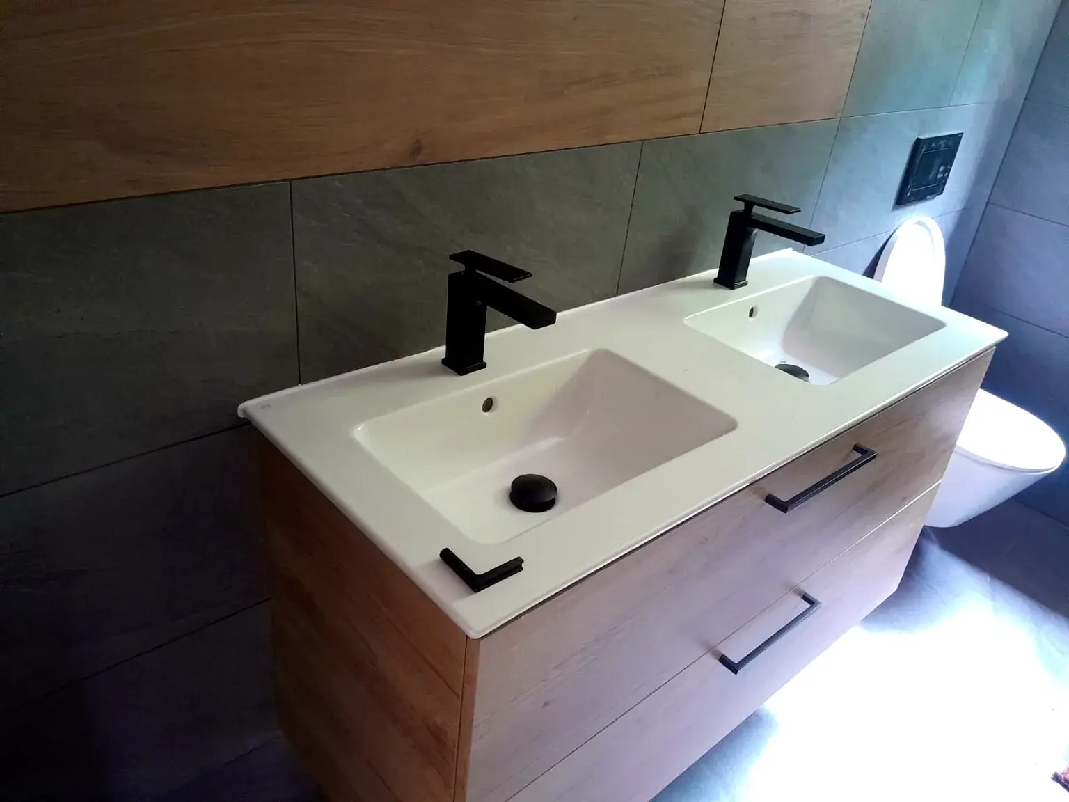 Meuble double vasque bois clair avec robinetterie noire sur carrelage gris.
