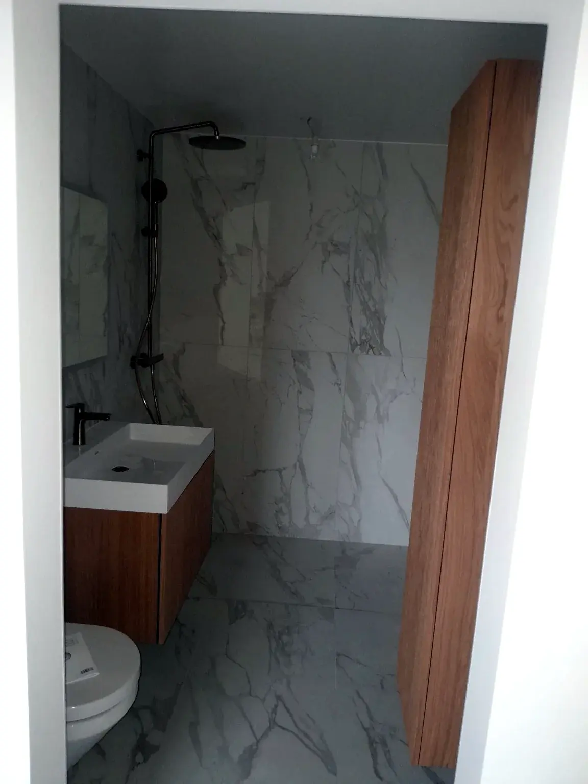 Salle de bains compacte avec revêtement effet marbre, meuble bois et douche.