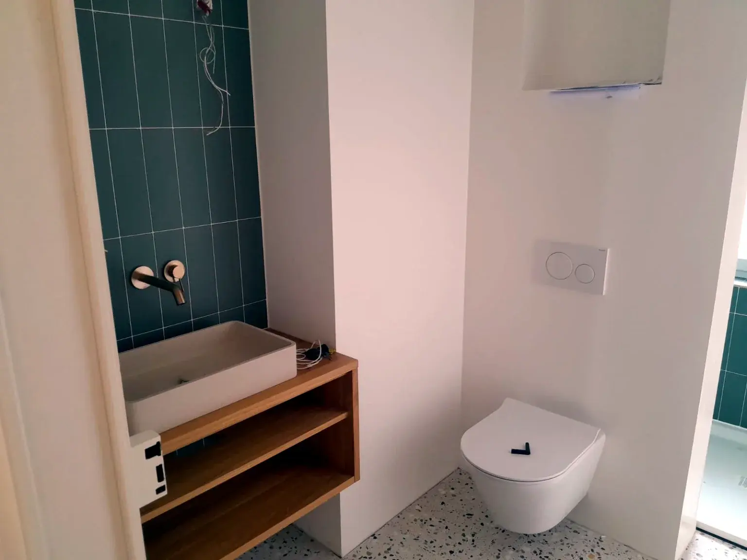 Petit espace sanitaire avec vasque sur meuble bois, robinetterie murale et carrelage vert.