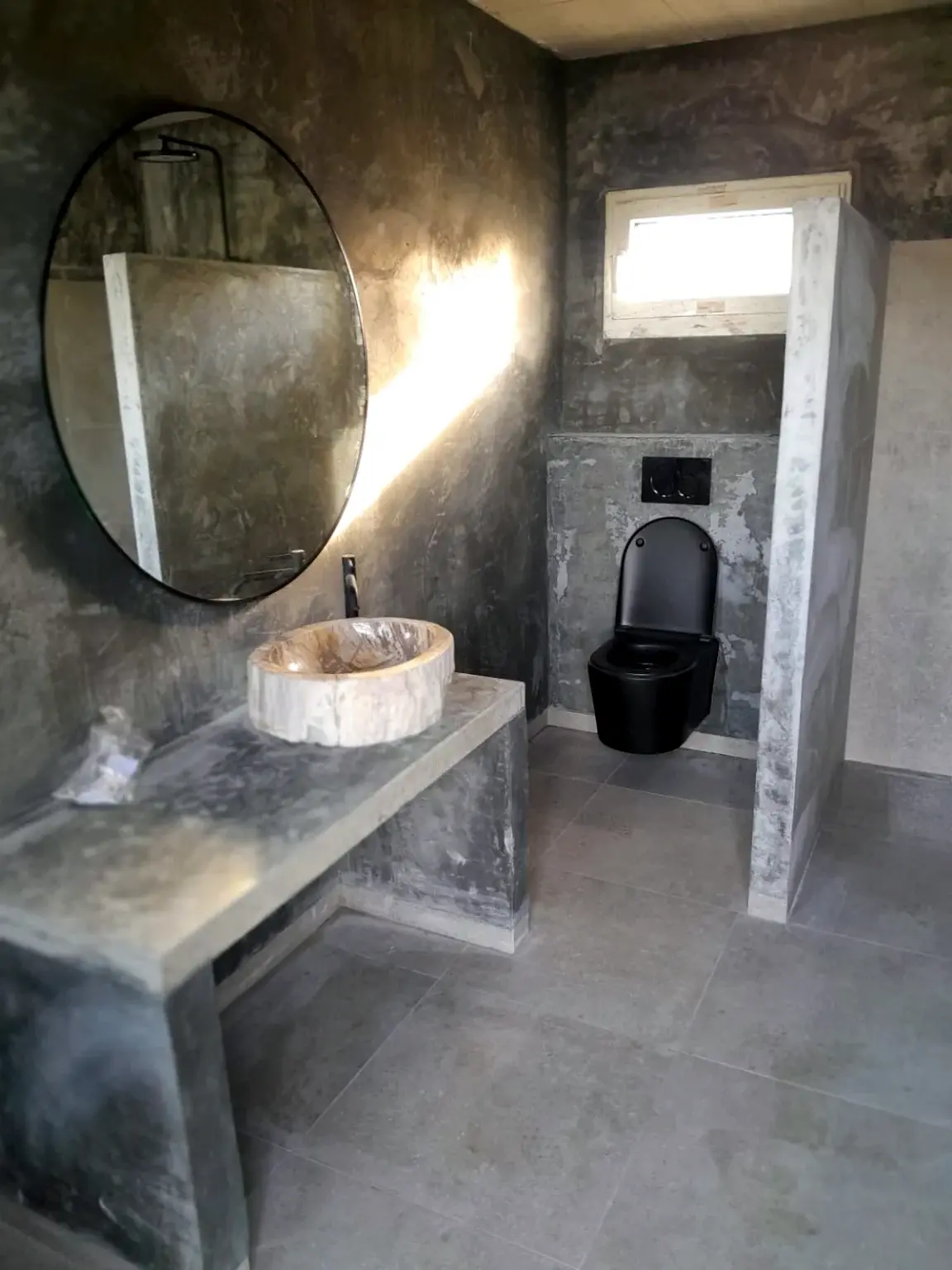 Salle de bains haut de gamme aux finitions minérales avec vasque en pierre, miroir rond et sanitaires noirs.