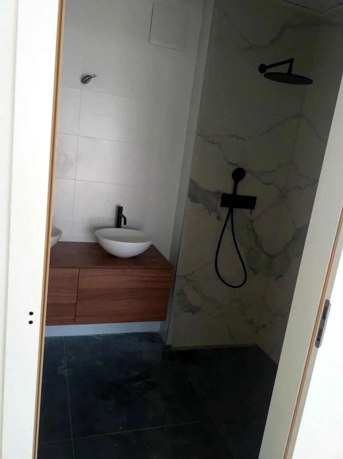 Salle de douche avec meuble vasque en bois, paroi effet marbre et robinetterie noire encastrée.