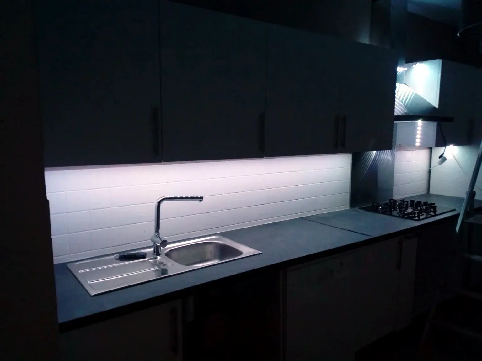 Cuisine contemporaine sombre avec éclairage sous meubles, évier inox et plaque de cuisson.