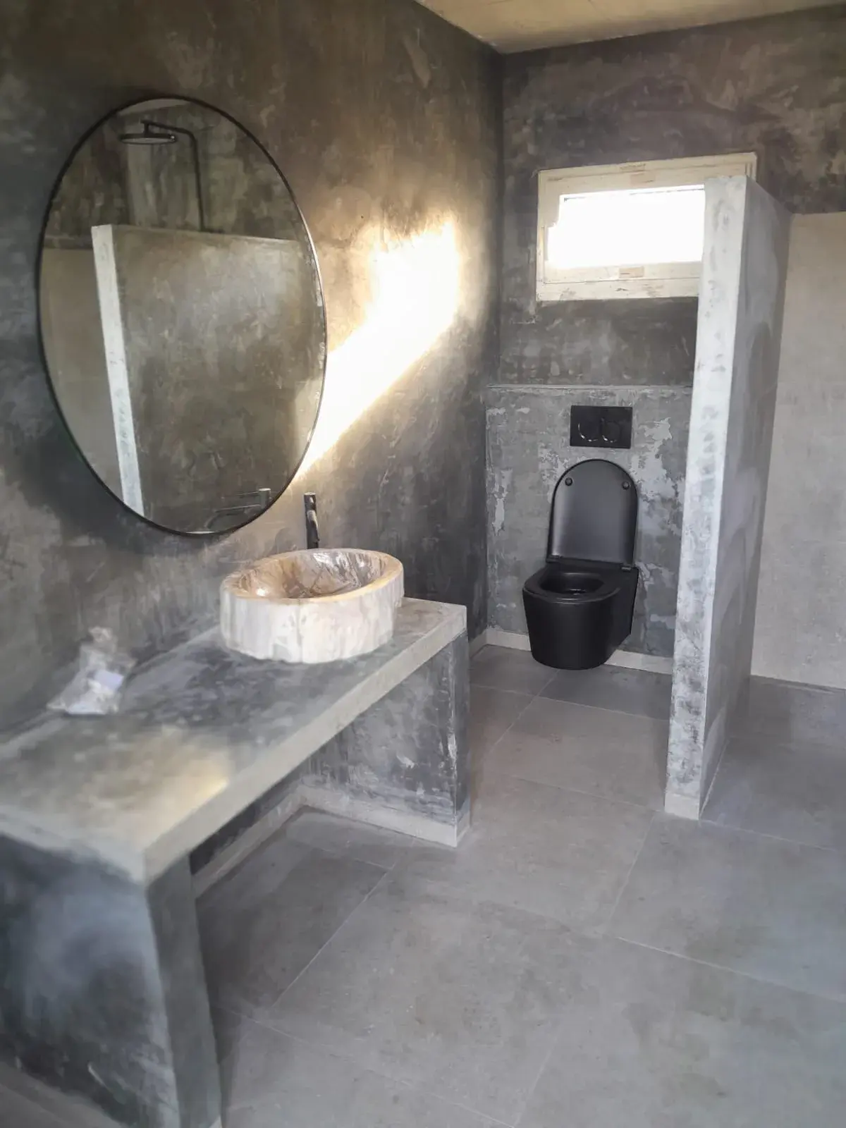 Vasque en pierre sur plan béton brut, miroir rond et WC noir, robinetterie noire.