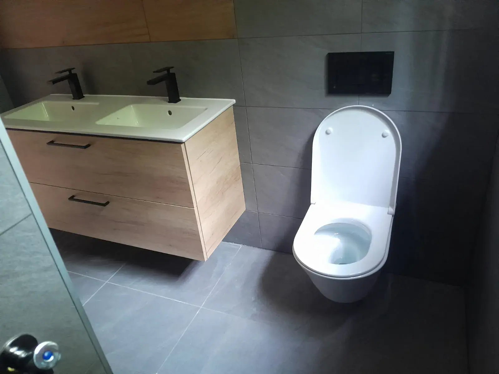 Double vasque en bois clair et WC suspendu avec plaque de commande noire.